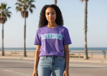 Load image into Gallery viewer, Person wearing a purple 'SUNKISSED' t-shirt by the beach with palm trees in the background