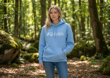 Load image into Gallery viewer, Woman wearing a blue hoodie with a logo in a forest setting
