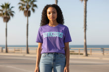 Load image into Gallery viewer, Person wearing a purple &#39;SUNKISSED&#39; t-shirt by the beach with palm trees in the background
